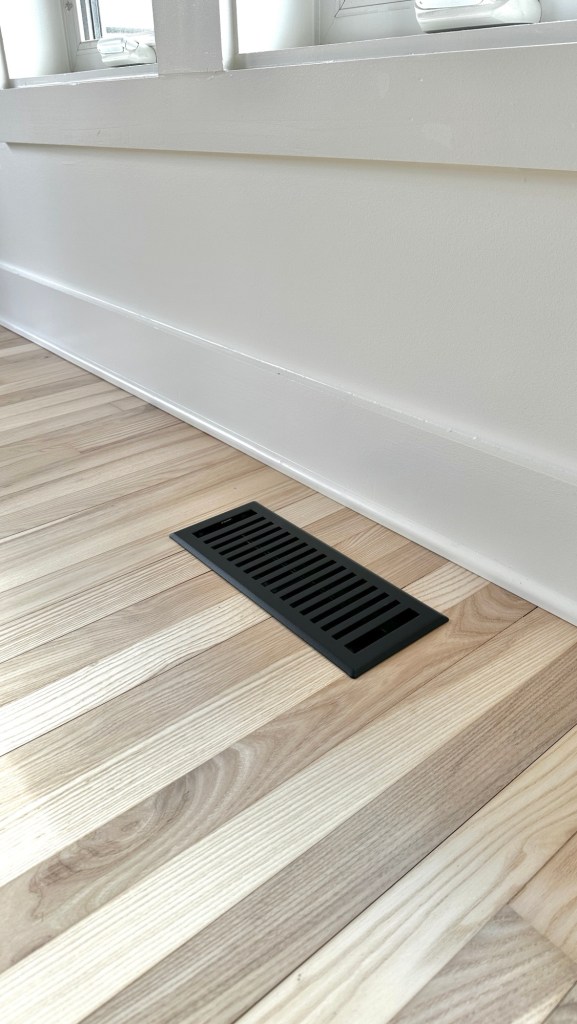 Close-up of a black floor vent grate installed on light wood flooring, with white walls and trim in the background.