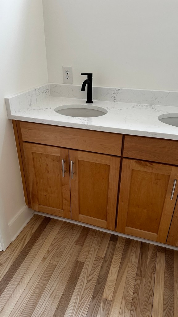A modern bathroom vanity with a marble countertop, two circular sinks, and wooden cabinetry beneath. The walls are painted white, and the floor features a wood pattern.