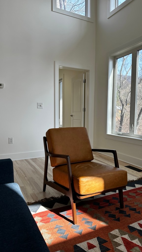 A cozy living space featuring a modern brown leather chair, a patterned rug, large windows allowing natural light, and a minimalist white wall design.