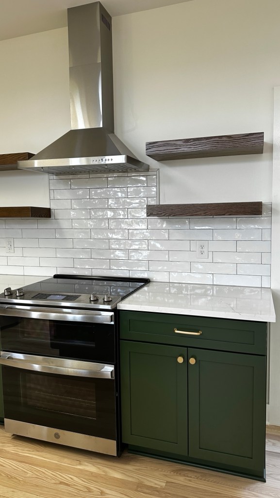 Modern kitchen with a green cabinet, stainless steel stove, and marble countertop, featuring white subway tiles and wooden shelves.