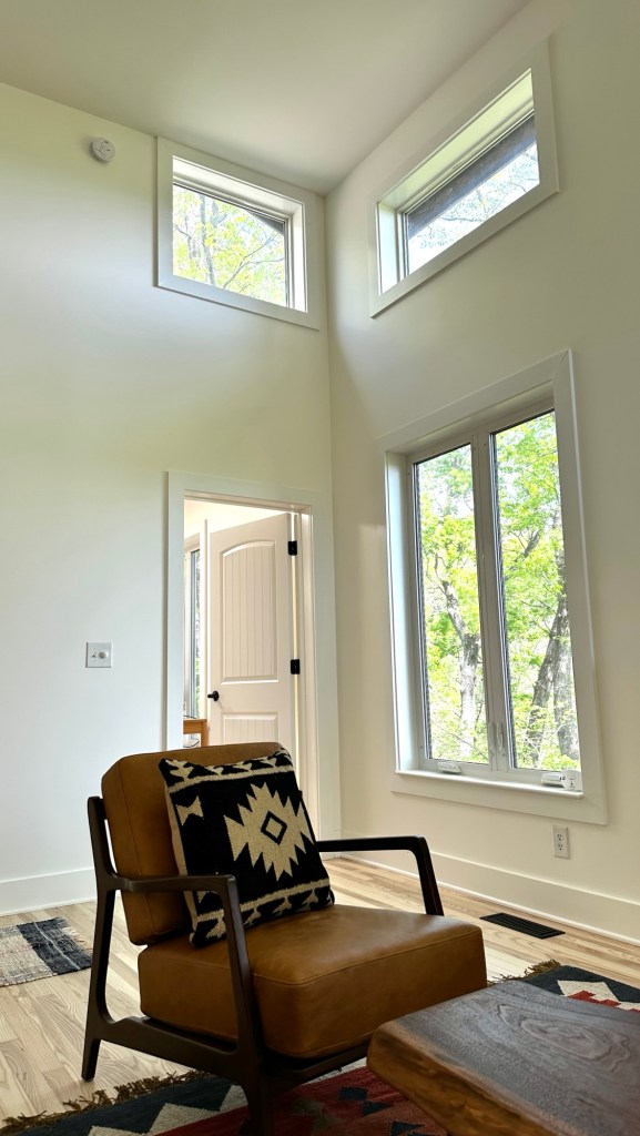 A cozy interior space featuring a modern cabin design with white walls, large windows allowing natural light, and a comfortable brown armchair adorned with a patterned pillow.